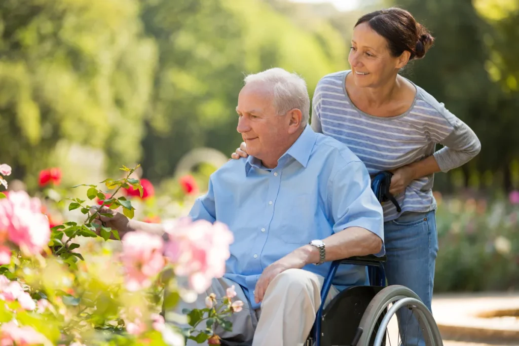 Elderly man in wheelchair gently holds blooming rose, guided by smiling caregiver in striped shirt. They are in a sunlit garden, creating a warm, joyful scene.